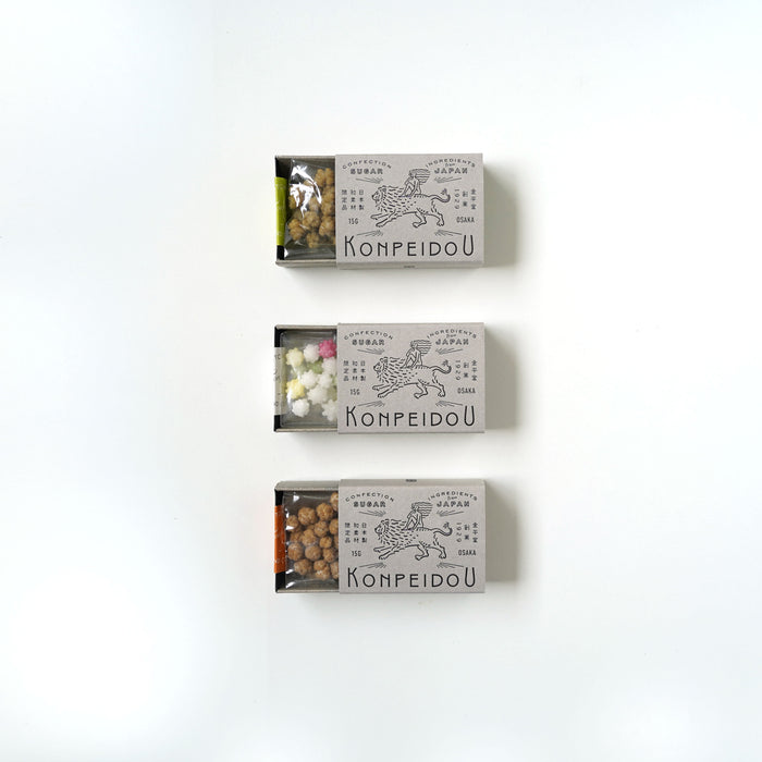 Three small rectangular boxes of Kompeidoó» sweets arranged vertically on a plain white background. The boxes are light gray with black line drawings and text, and the tops are slightly open to reveal the colorful contents inside.