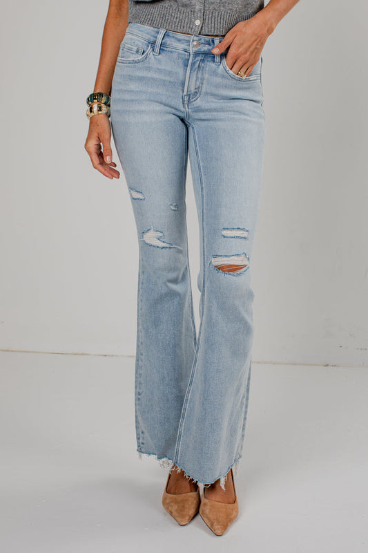 A person wearing light-wash blue distressed flare jeans with frayed hems, paired with tan pointed-toe heels and a gray top, standing against a plain light gray studio background.