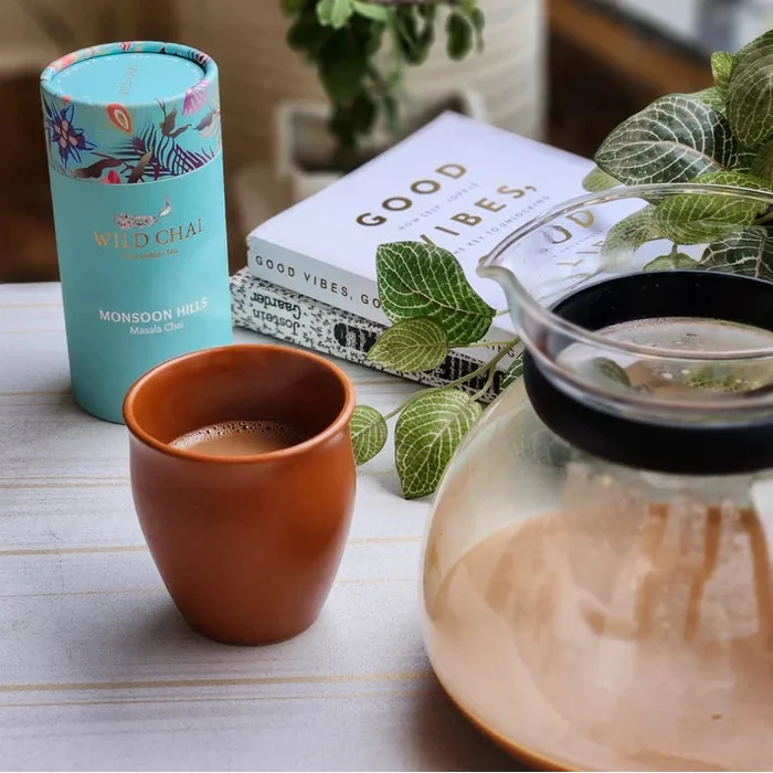 Tea packaging, cup, and glass teapot on a table with plants and books in the background