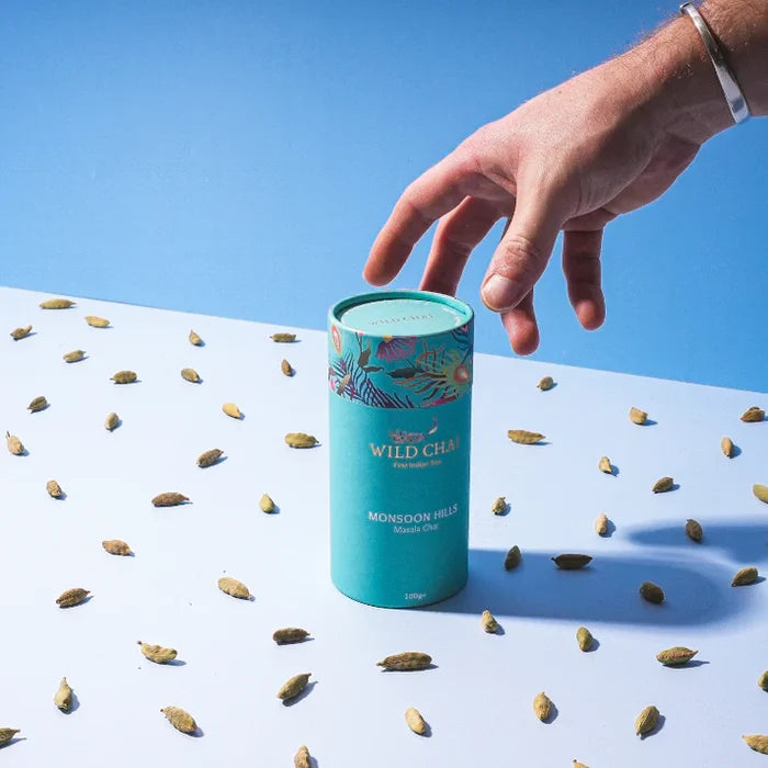 Hand reaching for a Wild Chai tea canister on a white surface with scattered tea leaves against a blue sky.