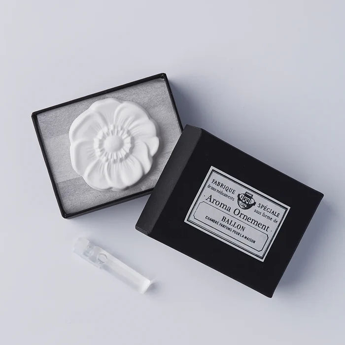 A white oil diffuser in the shape of an anemone, displayed in an open black box with a label, alongside a small clear vial.
