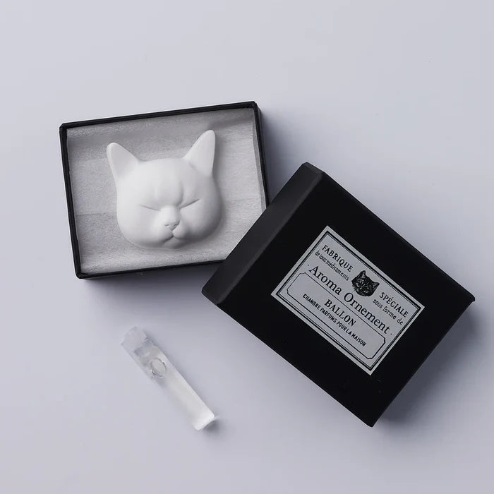 A white oil diffuser in the shape of a cat's head, displayed inside an open black box with a label. A small white stick, possibly for incense, is also shown.