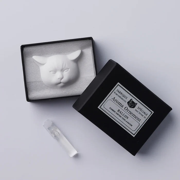 A white oil diffuser in the shape of a cat's head, displayed inside an open black box with a label. A diffuser refill is also visible beside the box.