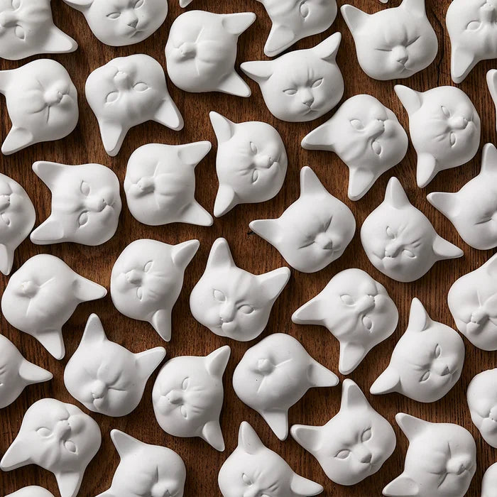 Pattern of white cat-face decorative objects arranged on a dark wooden background, featuring sculpted feline expressions in a minimalist modern product display.
