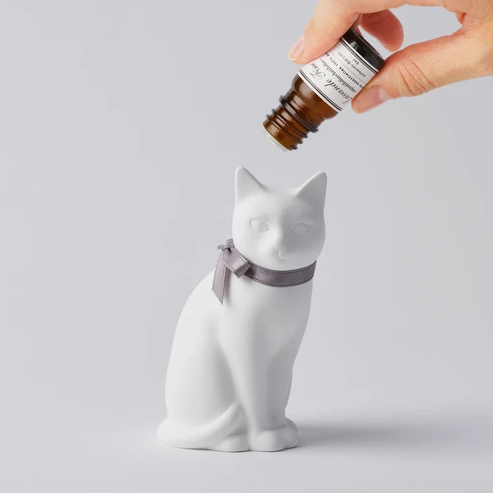 Hand applying fragrance oil to a white cat-shaped aroma ornament with a gray ribbon collar, shown on a clean white background in a minimalist home fragrance product scene.