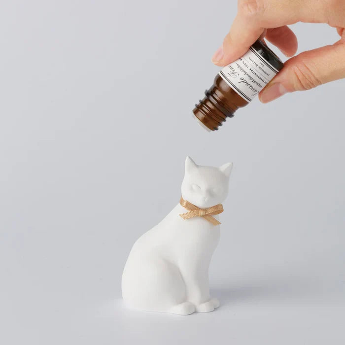 Hand applying fragrance oil to a white cat-shaped aroma ornament with a gold ribbon, shown on a clean white background in a minimalist home fragrance product scene.