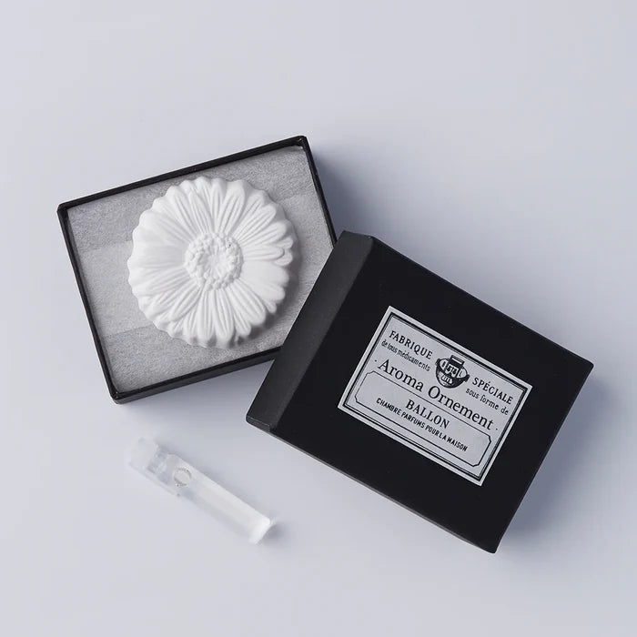 A white, flower-shaped oil diffuser placed inside an open black box with a clear glass vial beside it, on a grey background.