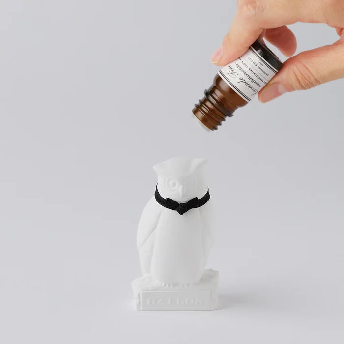 Hand applying fragrance oil to a white owl-shaped aroma ornament with a black ribbon collar, shown on a clean white background in a minimalist home fragrance product scene.