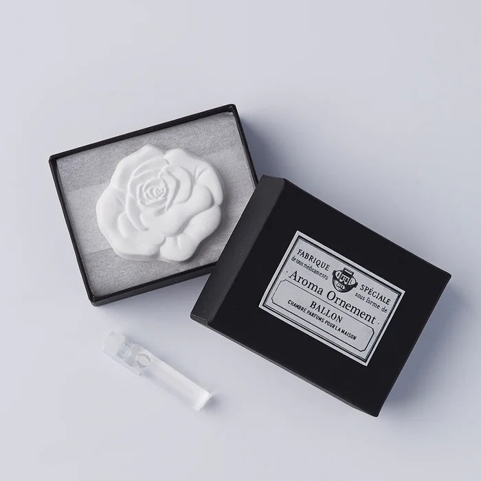 A white rose-shaped oil diffuser placed inside an open black box with a gray inner lining. A small spray bottle is also visible next to the diffuser.