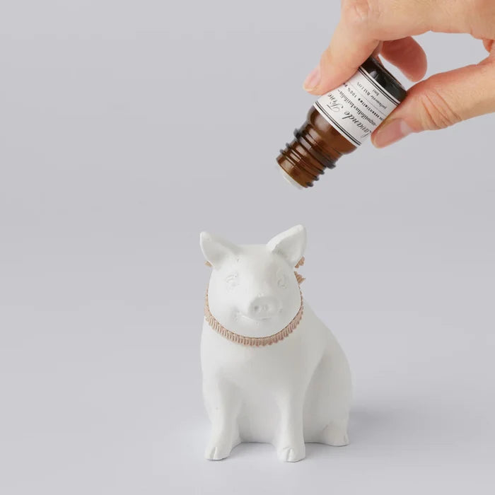 Hand applying fragrance oil to a white pig-shaped aroma ornament with a ribbon collar, shown on a clean white background in a minimalist home fragrance product scene.