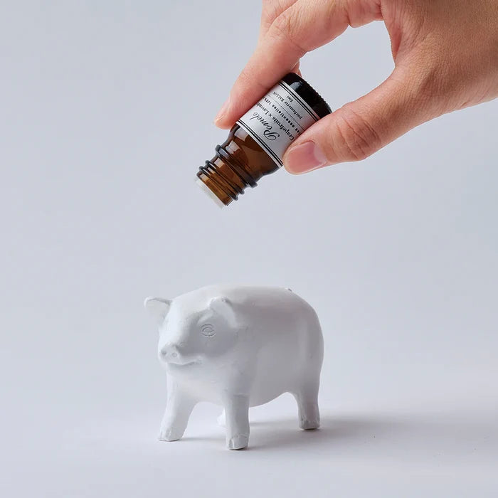 Hand applying fragrance oil to a white pig-shaped aroma ornament, shown on a clean white background in a minimalist home fragrance product scene.