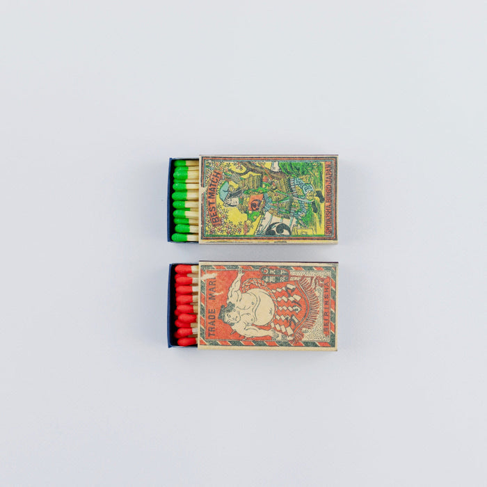 Two open Japanese matchboxes stacked vertically on a white background, showing colorful vintage illustrations on the covers. The top box reveals green-tipped matches and features a traditional warrior scene, while the bottom box shows red-tipped matches with a sumo wrestler illustration.