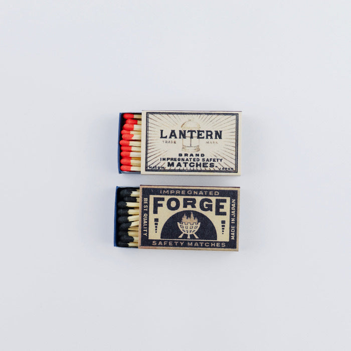 Two open vintage matchboxes stacked vertically on a white background. The top box is labeled “Lantern” and shows red-tipped matches, while the bottom box is labeled “Forge” with a bold black design and black-tipped matches, both featuring classic safety match branding.