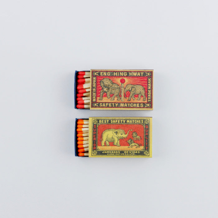 Two open vintage matchboxes arranged vertically on a white background. The top box features a red-and-gold label with elephants and red-tipped matches, while the bottom box shows orange-tipped matches and a colorful illustration of an elephant and handler, both in classic safety match packaging.
