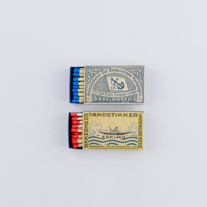 Two open vintage matchboxes stacked vertically on a white background. The top box shows blue-tipped matches and a blue-and-cream label with nautical imagery and Scandinavian-style text, while the bottom box displays red-tipped matches and a yellow label featuring an Arctic scene with waves, mountains, and retro typography.