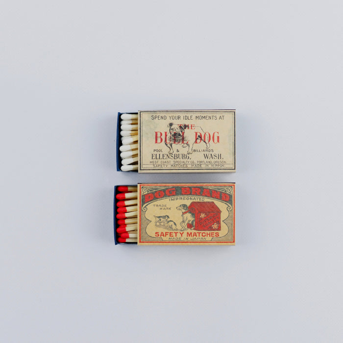 Two open vintage matchboxes stacked vertically on a white background. The top box shows white-tipped matches and a cream label with a bulldog illustration and retro text, while the bottom box features red-tipped matches and a red-and-tan “Dog Brand” safety matches design.