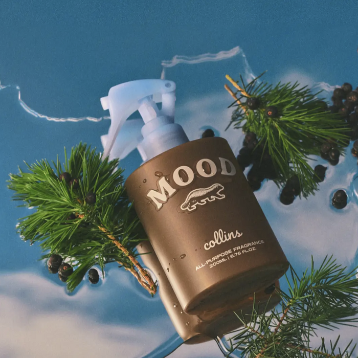 A bronze-colored room spray bottle labeled 'MOOD collins' with Scottish Juniper branding, displayed against a blue sky and foliage backdrop, suggesting a connection to nature.