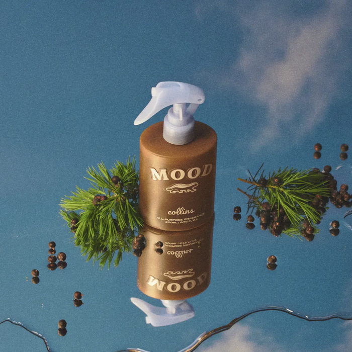 Mood all-purpose fragrance spray bottle reflected on a glossy blue surface with juniper sprigs and berries, artistic product styling for a fresh botanical home scent.