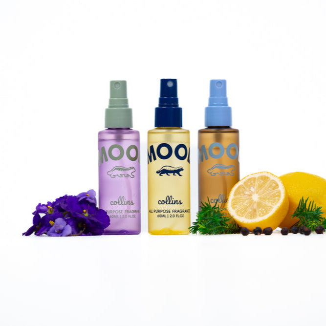 Three Mood Water all-purpose fragrance sprays in purple, yellow, and amber bottles displayed with violet flowers, lemon slices, juniper berries, and greenery on a clean white background.