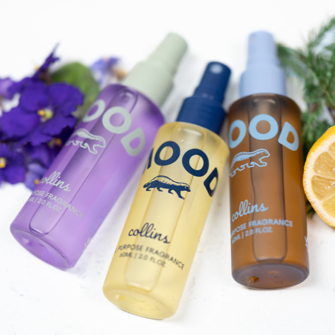 Close-up of three Mood Water all-purpose fragrance sprays in purple, yellow, and amber bottles, styled with violet flowers, lemon, and greenery for a fresh lifestyle product display.