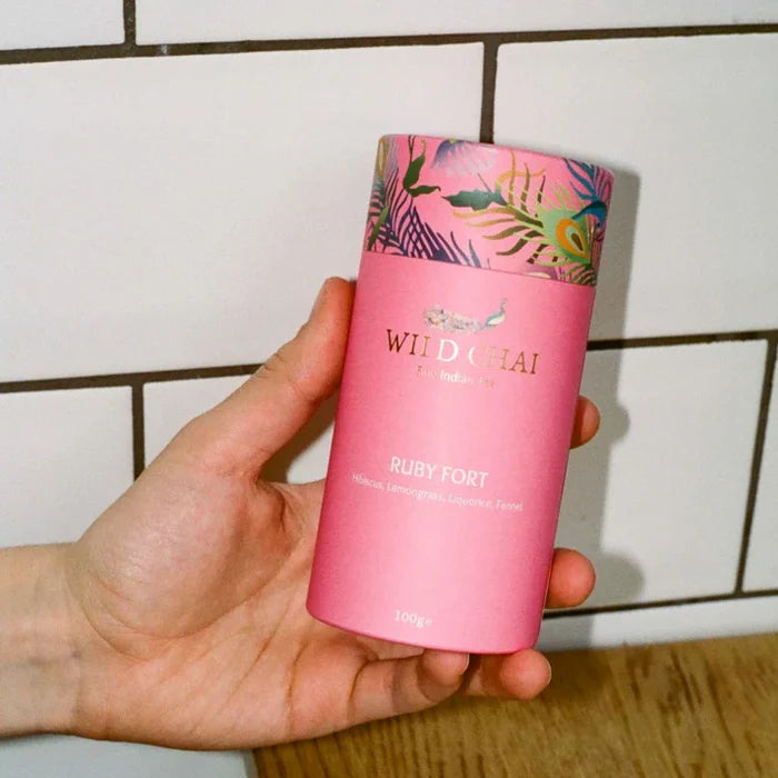 Hand holding a pink 'Wild Chai' container with a colorful lid against a tiled wall background.