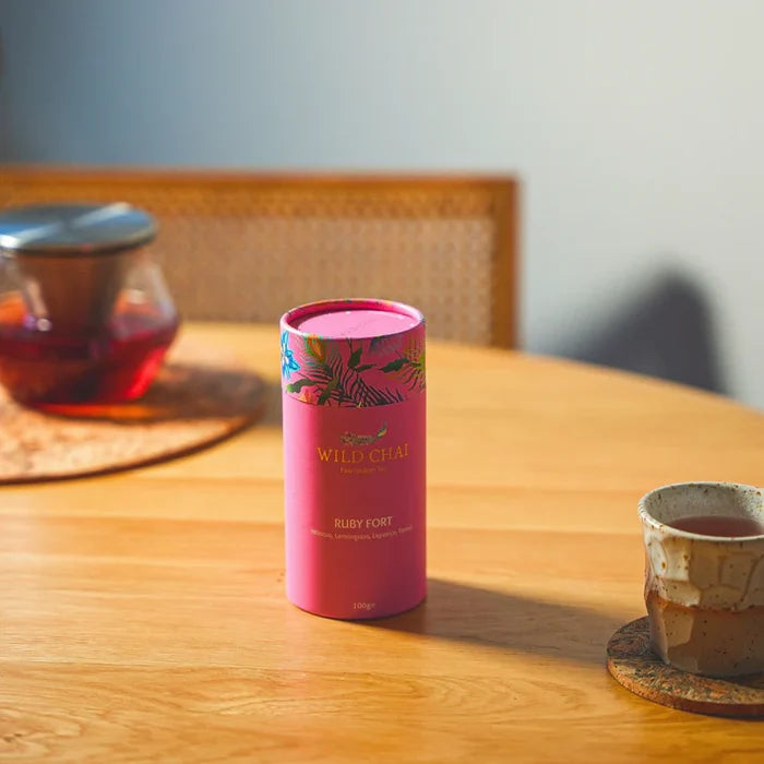 Pink cylindrical tea canister labeled 'Wild Chai' on a wooden table with a teacup and teapot in the background.