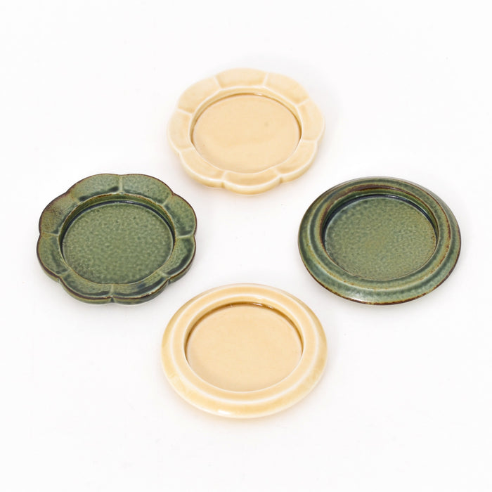 Four small ceramic dishes arranged on a white background, featuring round and scalloped designs in earthy green and warm cream glazes, suitable as match strikers or small trays.