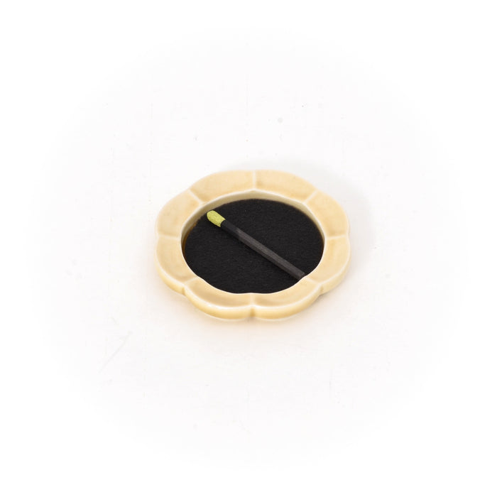 A small scalloped ceramic dish on a white background holding a round black striker pad, with a single incense match resting diagonally across the center.