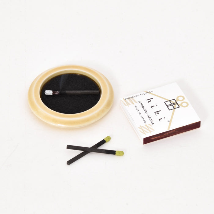 A hibi incense match set on a white background, featuring a cream ceramic dish with a black striker pad and one match resting inside, two loose incense matches in front, and a hibi 10 Minutes Aroma matchbox labeled “Japanese cypress” placed to the side.