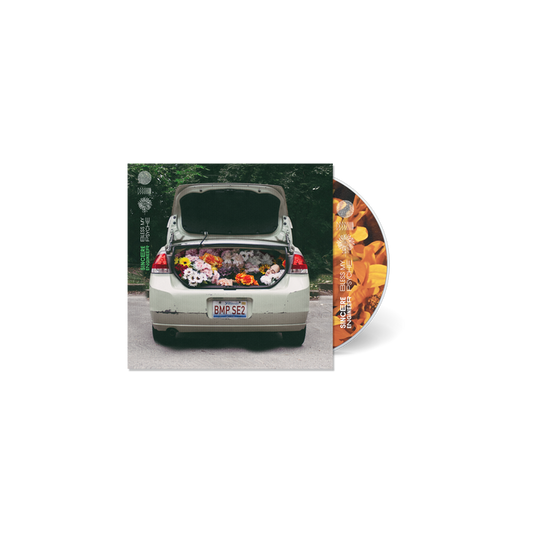 A car with an open trunk filled with colorful flowers is parked on a street. To the right, a Sincere Engineer CD for the Bless My Psyche album, featuring an orange floral design, is partially visible next to the album cover.