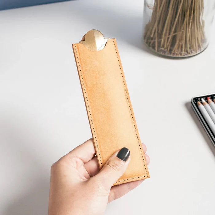 Hand holding a tan leather pencil case on a light surface with a jar of pencils in the background.