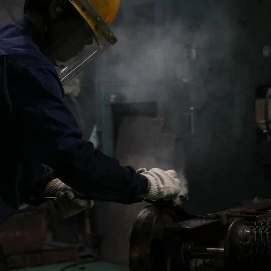 A worker in a dark blue uniform, white gloves, and a yellow hard hat handles a glowing orange-red piece of molten metal in an industrial setting with smoke and machinery.