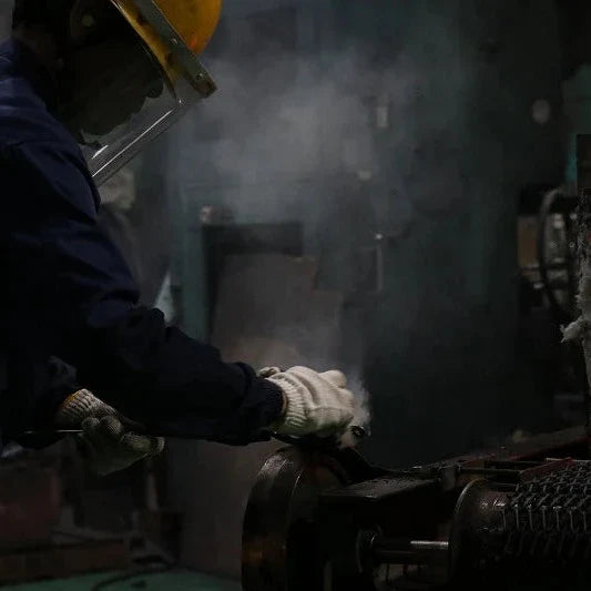 A worker in a dark blue uniform, white gloves, and a yellow hard hat handles a glowing orange-red piece of molten metal in an industrial setting with smoke and machinery.
