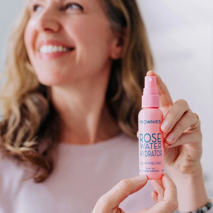 A smiling model showcasing her Frownies rose water hydrator spray. Her face and background are slightly blurred, making the product the focal point.