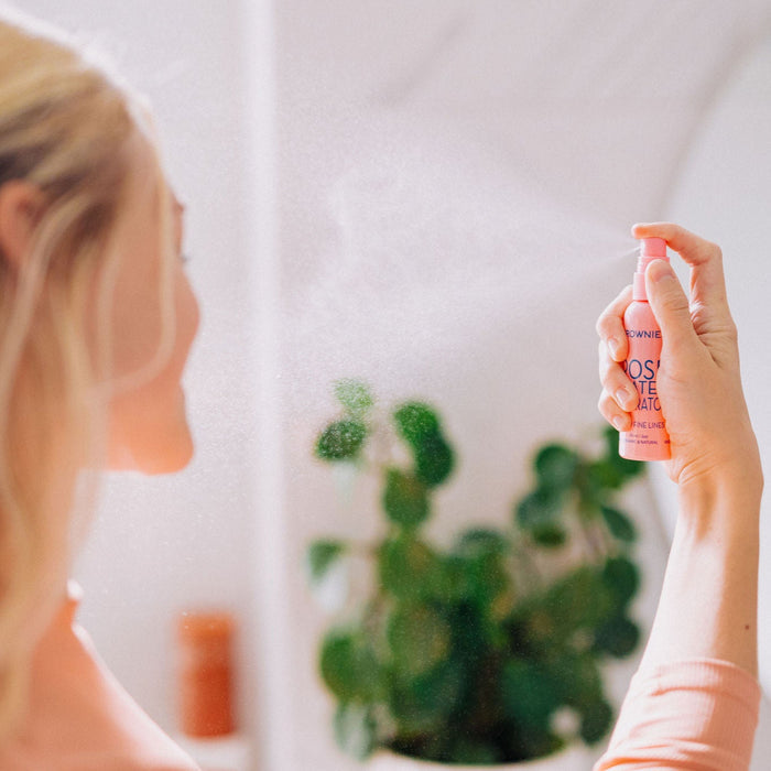 A woman with blonde hair shown spraying Frownies rose water hydrator spray onto her face.