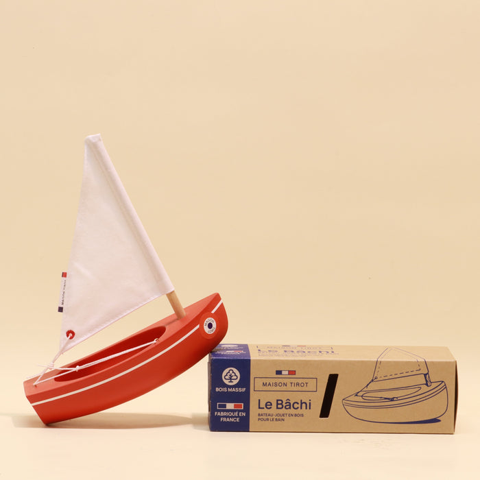 Red toy sailboat with white sail next to its packaging on a beige background