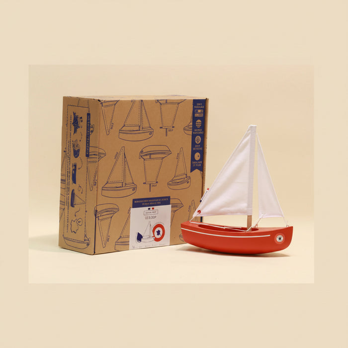 Red toy sailboat with white sail next to its packaging on a beige background