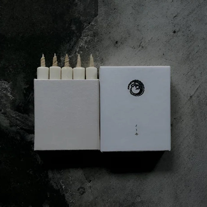 Set of five white mini taper candles in a minimalist white presentation box with matching sleeve, displayed on a dark textured surface in a modern product presentation.