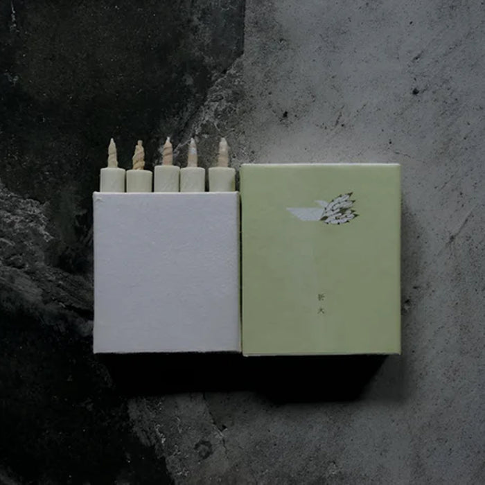 Set of five pale green mini taper candles in a minimalist white presentation box with matching sage green sleeve, displayed on a dark textured surface in a modern product presentation.