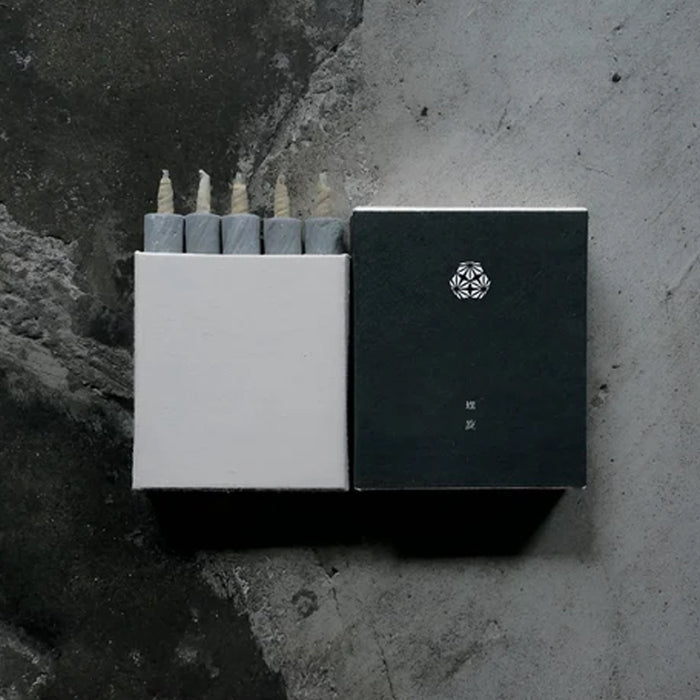 Set of five gray mini taper candles in a minimalist white presentation box with matching dark sleeve, displayed on a dark textured surface in a modern product presentation.