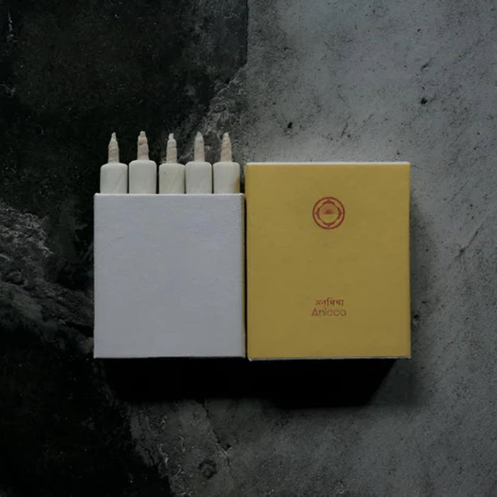 Set of five white mini taper candles in a minimalist white presentation box with matching mustard yellow sleeve, displayed on a dark textured surface in a modern product presentation.