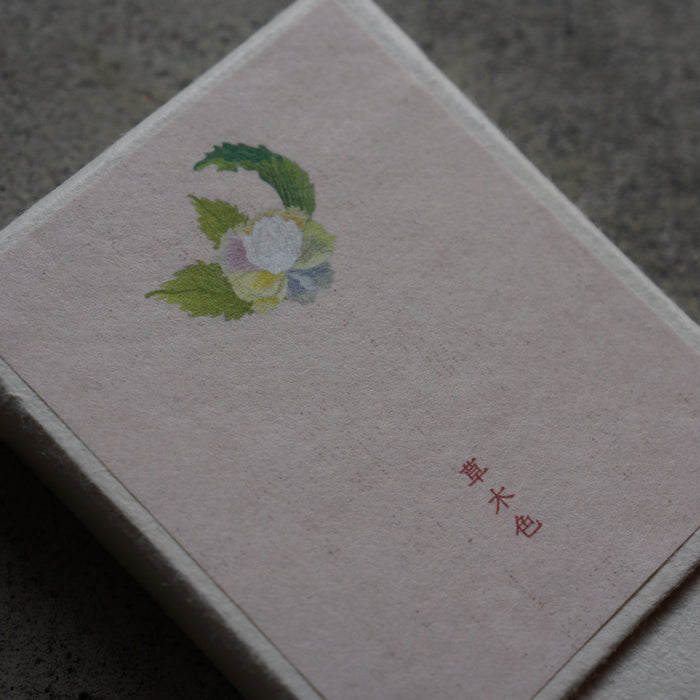 A square, textured light pink book or notebook with a small watercolor flower illustration and red Japanese characters on the cover, resting on a dark gray concrete surface.