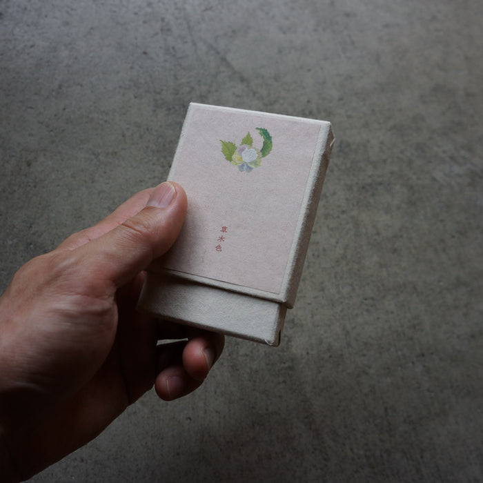 A hand holding a small square box with a light beige cover and a small green and white floral illustration near the top, set against a textured gray concrete background.