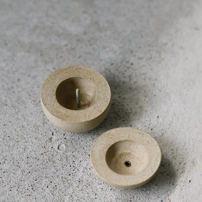 Minimalist round candle holder in a natural stone finish shown in two parts, displayed on a textured concrete surface in a modern product presentation.