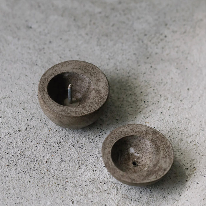 Minimalist round candle holder in a dark stone finish shown in two parts, displayed on a textured concrete surface in a modern product presentation.