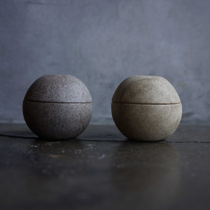 Set of two minimalist spherical candle holders in dark and light stone finishes, displayed on a dark tabletop against a moody gray background in a modern decor product presentation.