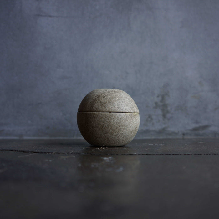 Minimalist spherical candle holder in a light stone finish, displayed on a dark tabletop against a moody gray background in a modern decor product presentation.