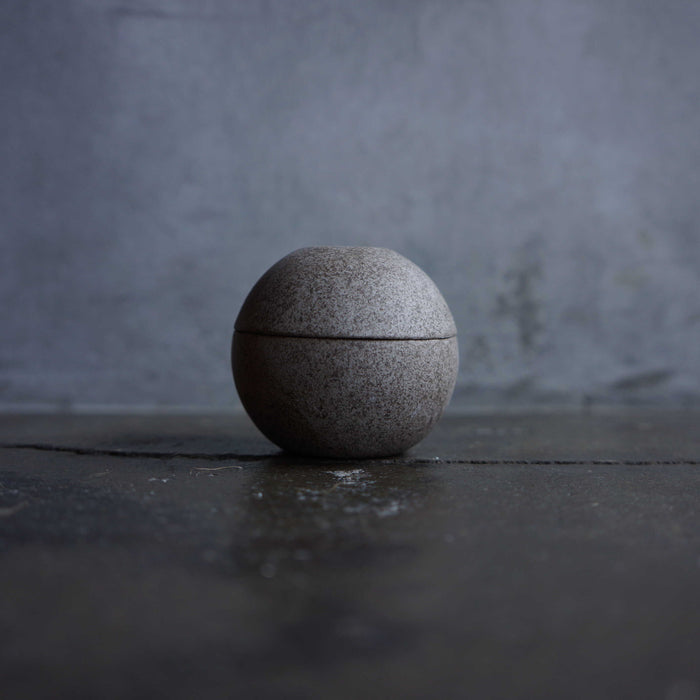 Minimalist spherical candle holder in a dark stone finish, displayed on a dark tabletop against a moody gray background in a modern decor product presentation.