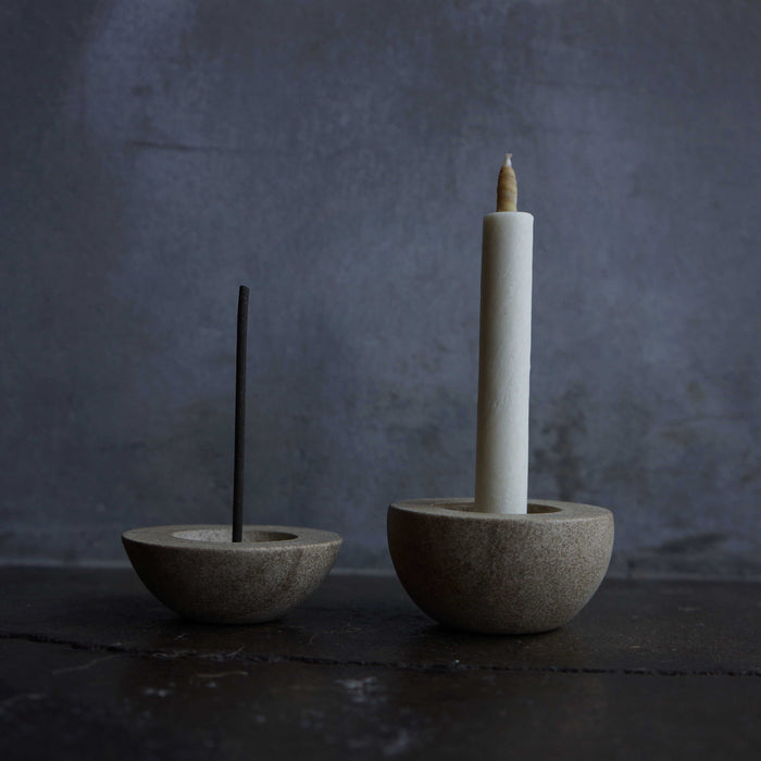 Minimalist stone holders for incense and taper candles, styled with a black incense stick and white candle on a dark tabletop against a moody gray background.