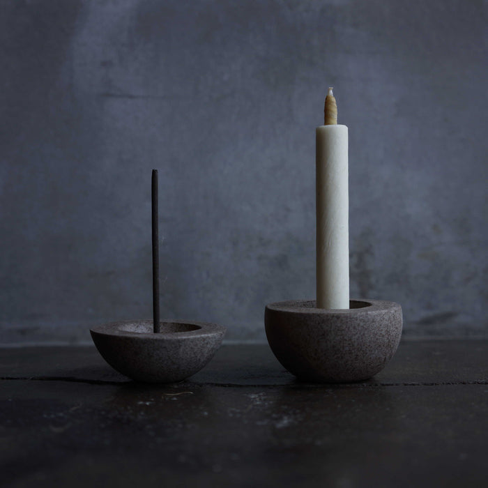 Minimalist dark stone holders for incense and taper candles, styled with a black incense stick and white candle on a dark tabletop against a moody gray background.